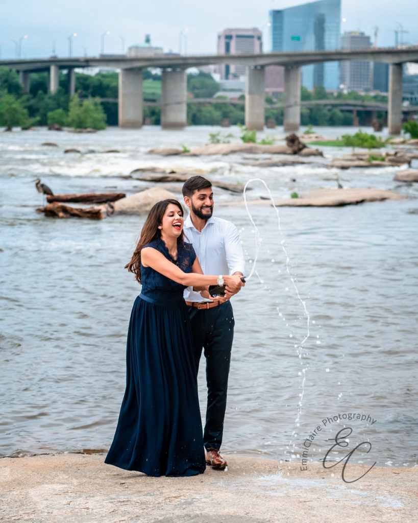 engaged couple pops a bottle of champagne and sprays it as they stand together beside a river