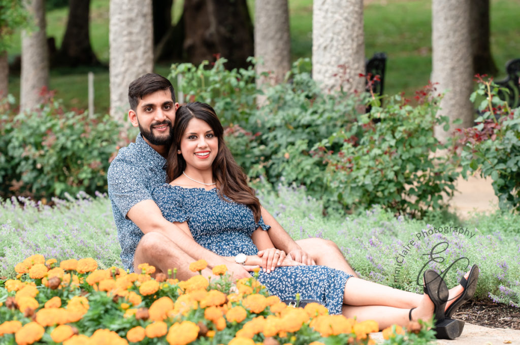woman in a blue dress sits down leaning against her fiance who is behind her on the grass in front of big yellow flowers