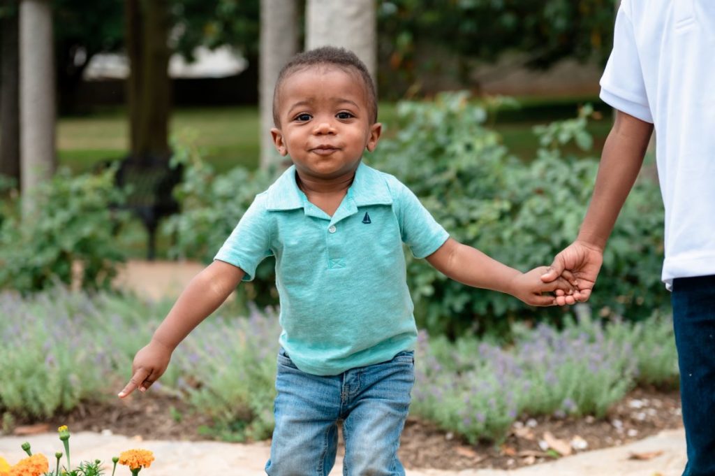 Small child in a teal collared shirt holding his older brother's hand as he points down to a yellow flower in an outdoor garden space