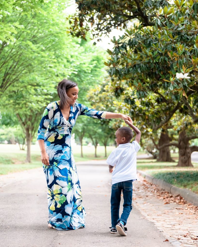 Mom dances and twirls in a floral blue dress with her young son outdoors amongst green trees