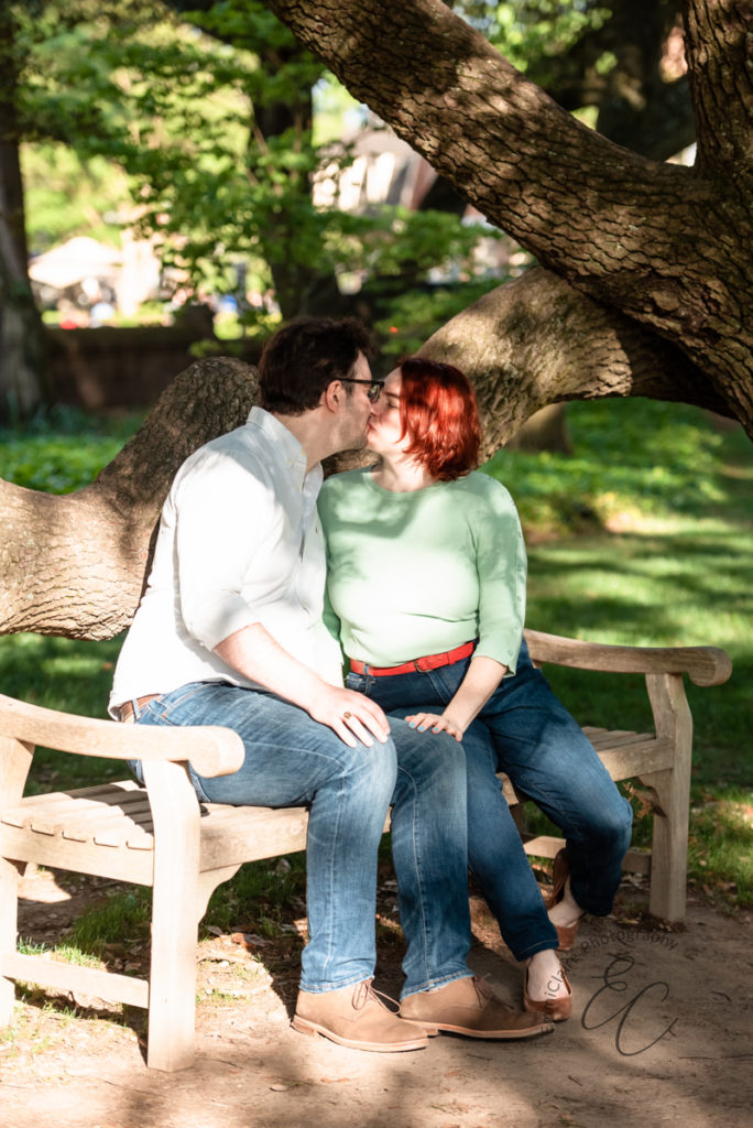 cute couple smooches under the shade of a large tree while sitting on a bench during their engagement photoshoot session