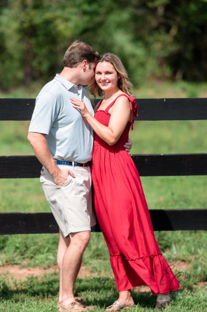 woman standing in a red dress next to a fence holding her partner and smiling after becoming engaged 