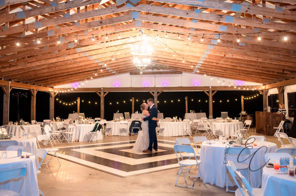 bride and groom share a dance on the dance floor before their guests arrive at the reception under a wooden-beamed space with white tables and a chandelier 