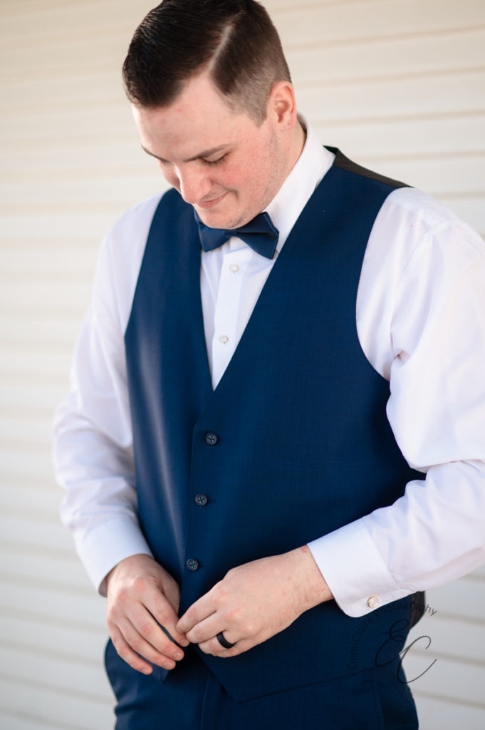 groom stands outside buttoning his blue vest and smiling as he awaits his pastoral wedding ceremony