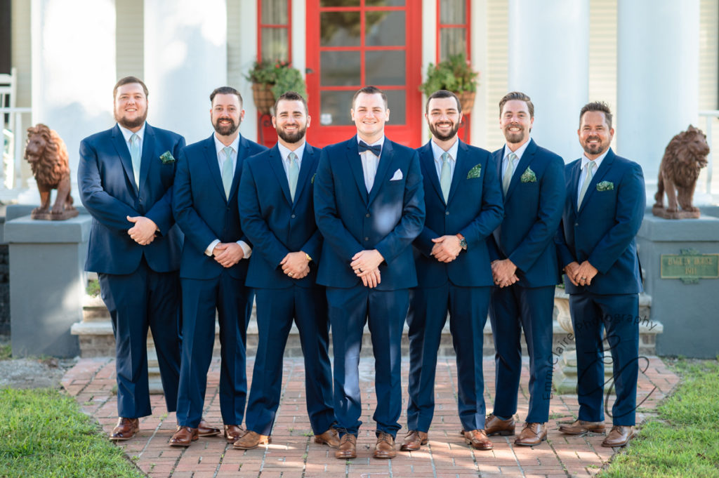 groom stands in the middle of his six groomsmen in front of their wedding property manor all wearing navy blue tuxedos