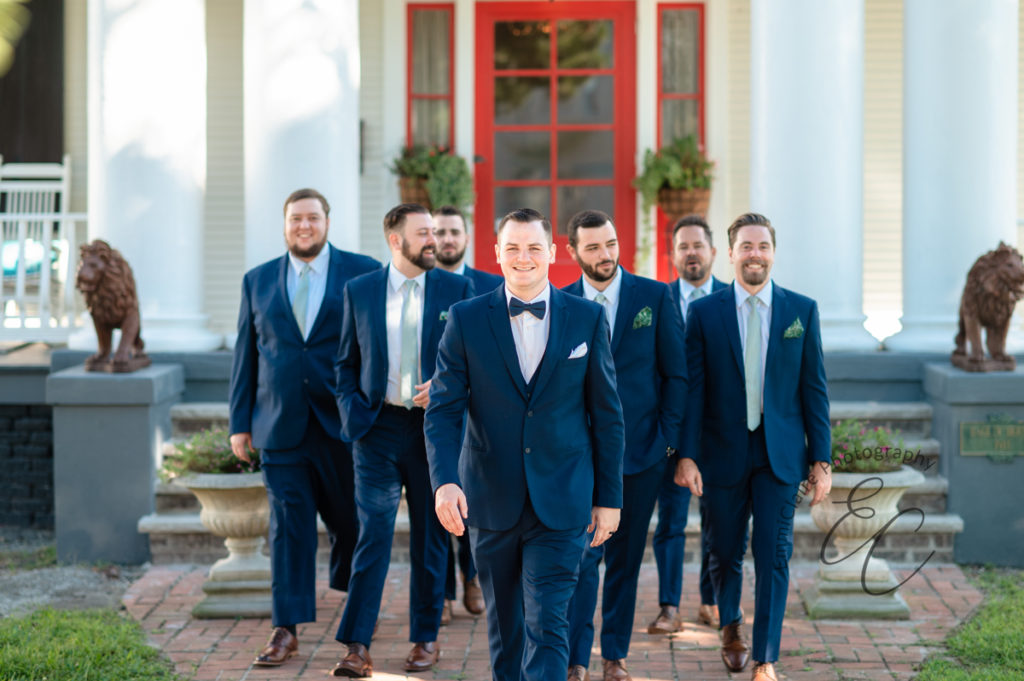 groom walks towards the camera smiling as his groomsmen trail behind him all wearing navy blue tuxedos out front of the wedding venue home