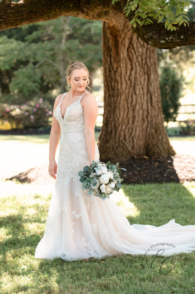 Bride standing in front of a large tree in her beaded, detailed wedding dress while holding her bouquet down at her side and looking towards the grass