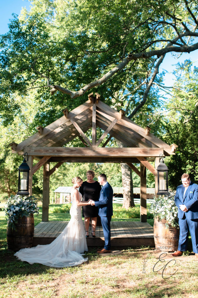 bride and groom standing in the sun outside facing one another holding their hands out in front of one another as they say "I do" at their pastoral-style wedding