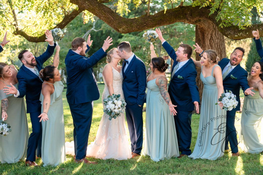 bride and groom share a kiss as their bridesmaids and groomsmen stand among them under a large tree cheering and celebrating