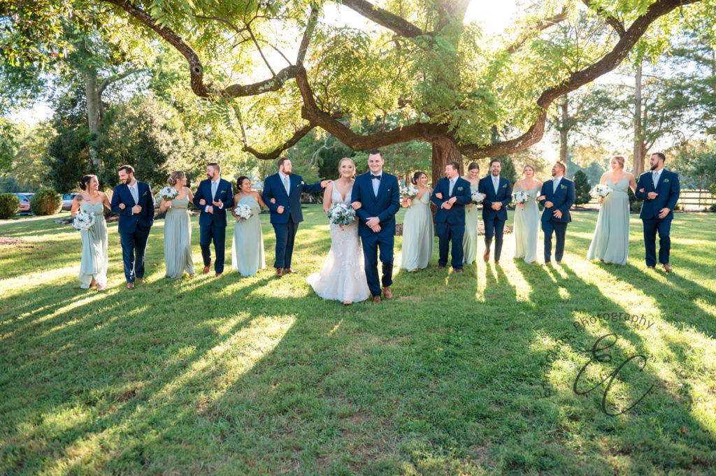bride and groom stand just slightly in front of their bridal party who are all lined up behind them smiling at the camera and enjoying the outdoors on their wedding day
