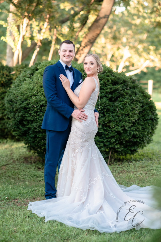 Bride and groom stand in front of a large green shrub facing the camera and smiling for their wedding photos they will cherish for a lifetime