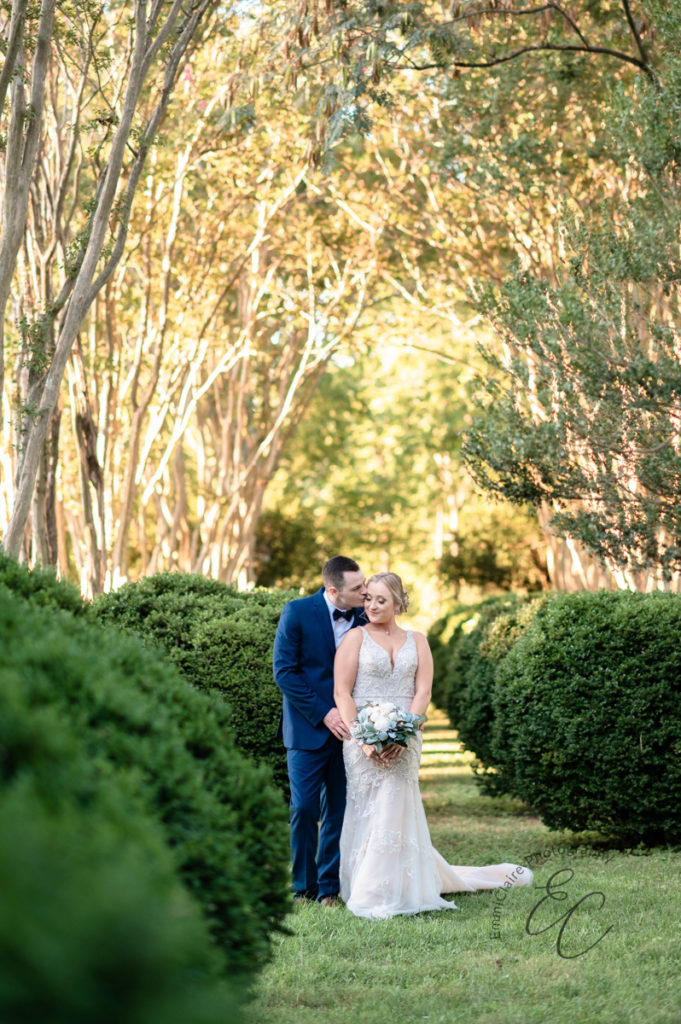 groom stands under tree shade with his bride following their beautiful outdoor ceremony and kisses her on the forehead