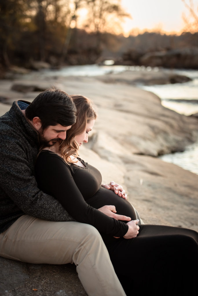 Expectant mother lays against her partner as they sit down together on the banks of a river at sunset for their maternity photos