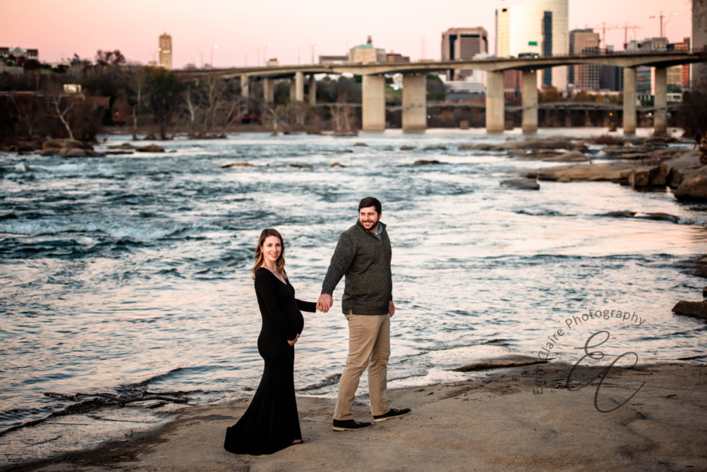 Parents who are expecting their first child stand by a river with the cityscape in the background during their maternity photoshoot