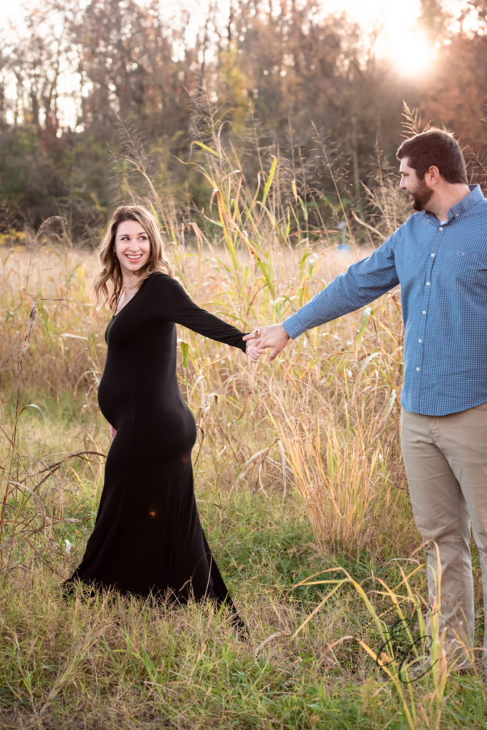 expectant parents stand together outside in the tall grass holding hands and smiling during their maternity photoshoot