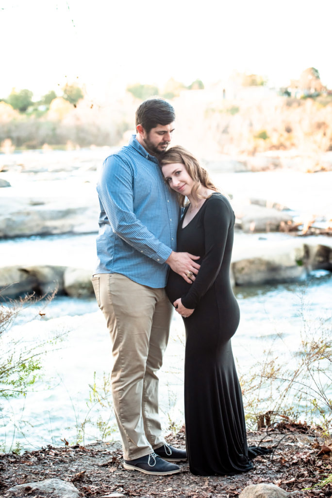 Expectant parents wrap their arms around mom's pregnant belly while standing in front of a flowing river 