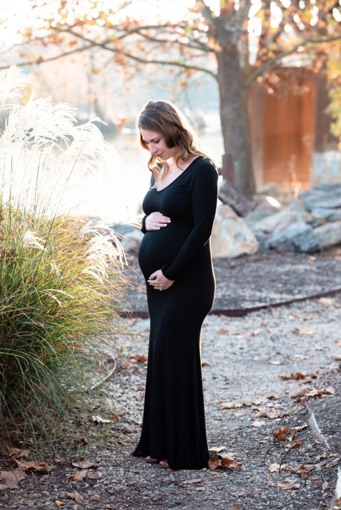 woman stands in a black dress embracing her pregnant belly with both hands while being captured by her photographer for every season of life