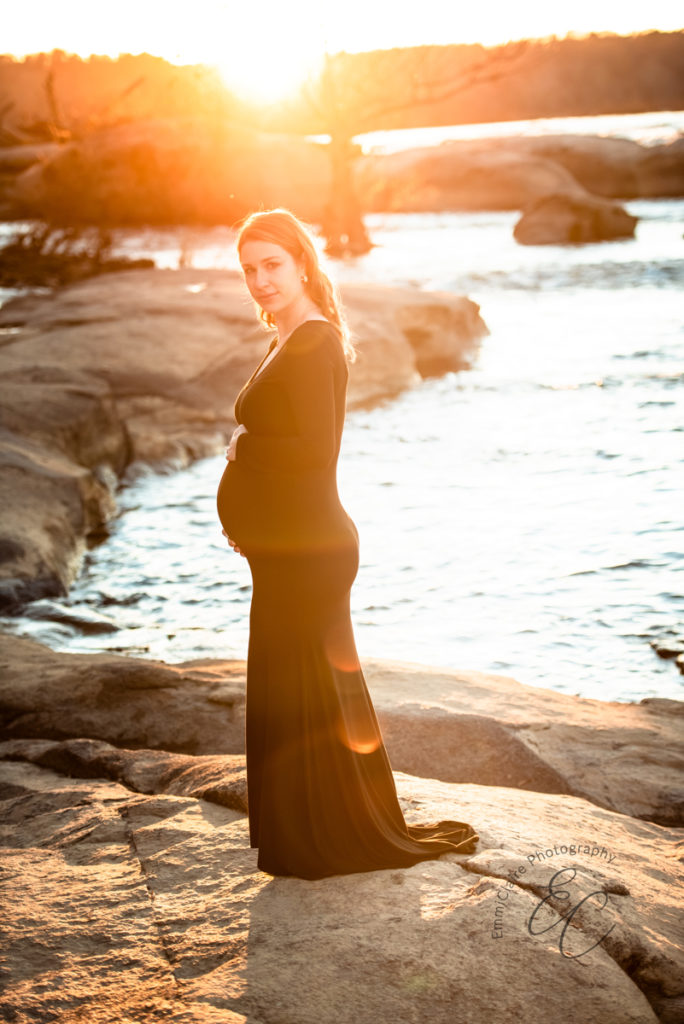 Pregnant woman stands by a flowing river at sunset embracing her pregnant belly with excitement to meet her child