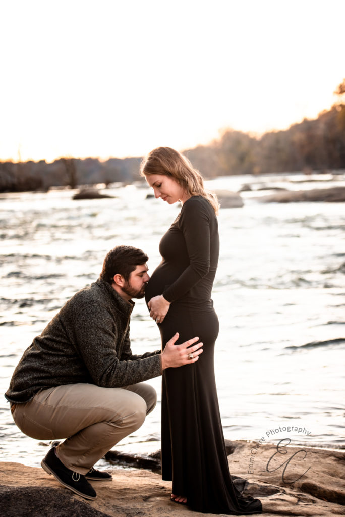Father-to-be kneeling down to kiss his partner's pregnant belly while she stands in front of a river in a beautiful long black dress
