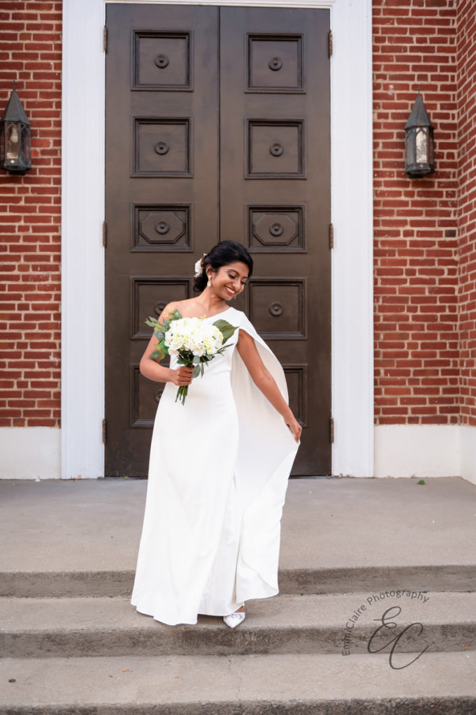 Beautiful bride stands in her one-shoulder white wedding grown holding her white bouquet in front of the church where her elopement ceremony will take place