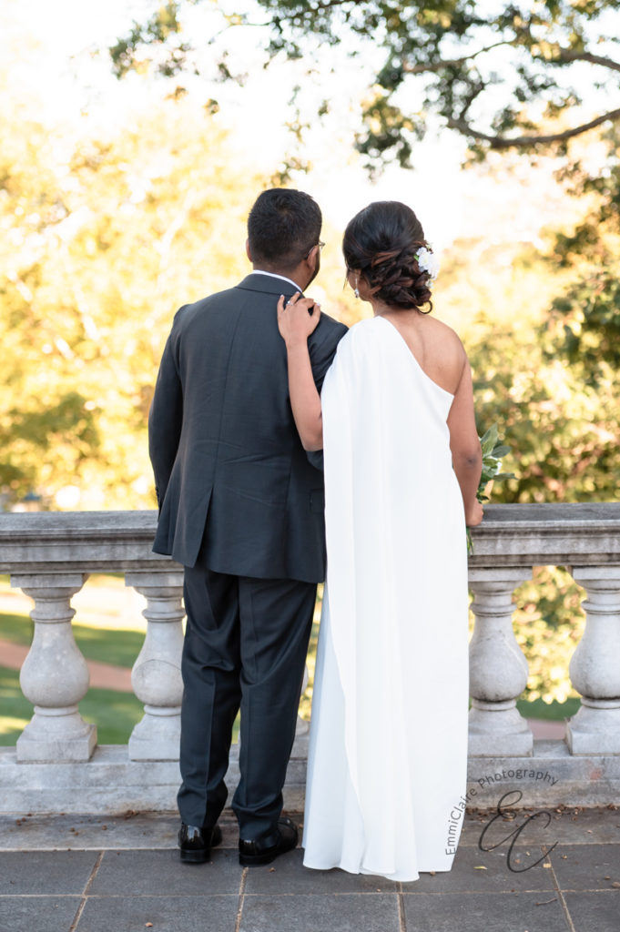 newlywed couple stands together with their backs to the camera facing out from a balcony towards a lush green space after their wedding ceremony