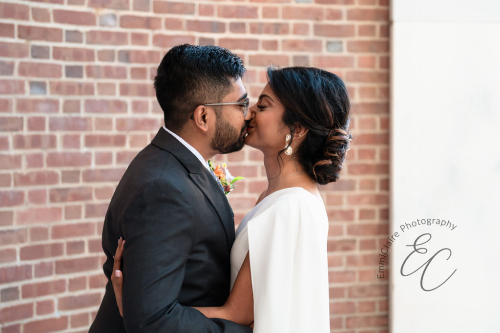 bride and groom standing in front of a brick wall hold ing onto one another and share a romantic kiss during their Charlottesville elopement 