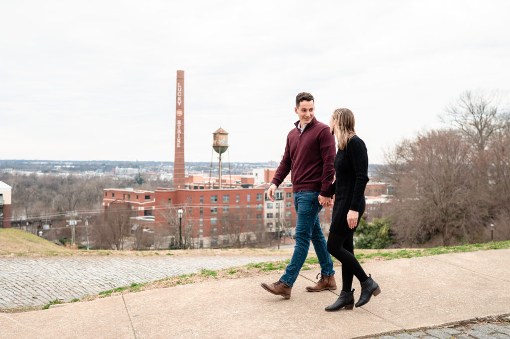 Caroline and Justin hold hands and look at one another as they take a walk along a Libby Hill Park path with the Lucky Strike building and water tower in the background. 