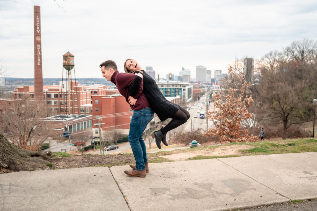 Justin lifts a laughing Caroline on his back with the Lucky Strike building and Richmond Skyline in the distance.