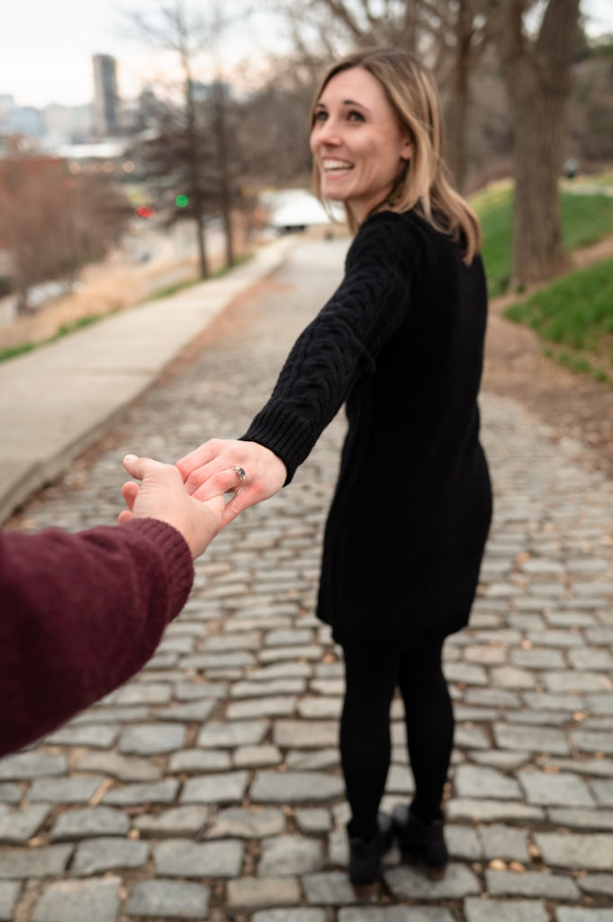 Caroline smiles as looks back at Justin (who is out of the frame) as she holds his hand. Bare February trees line a stone-paved street behind them. 