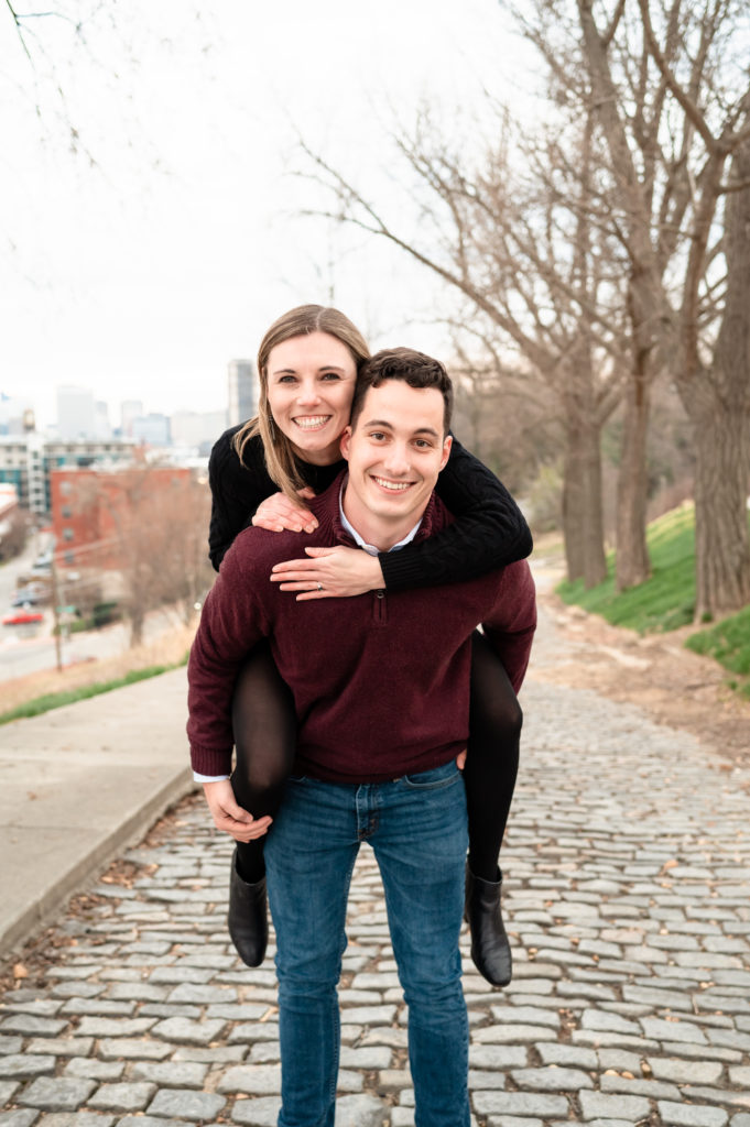 Justin gives Caroline a piggy back ride as the couple smiles directly at the camera along a stone street lined by bare winter trees. Behind them, the Richmond Skyline is in the distance.