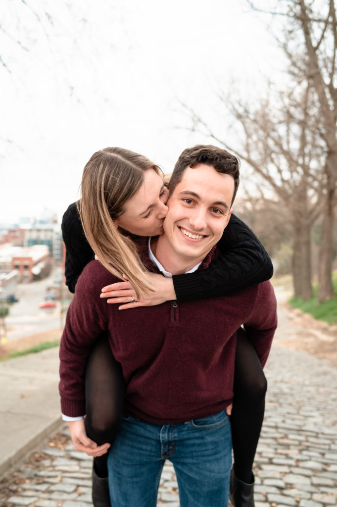 Justin gives Caroline a piggy back ride as he smiles directly at the camera while Caroline gives him a kiss on the cheek immediately following their proposal at Libby Hill.