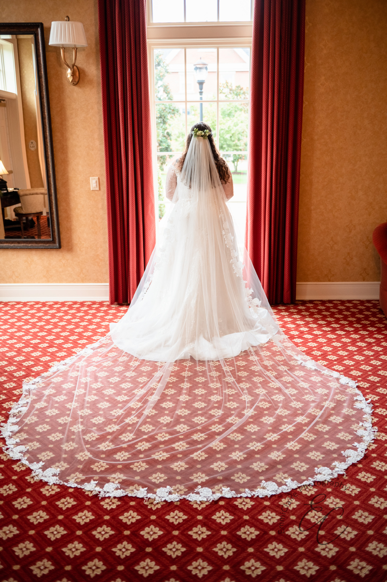 A color photo of the back of the bride's dress and veil as she stands in front of a floor to ceiling window.