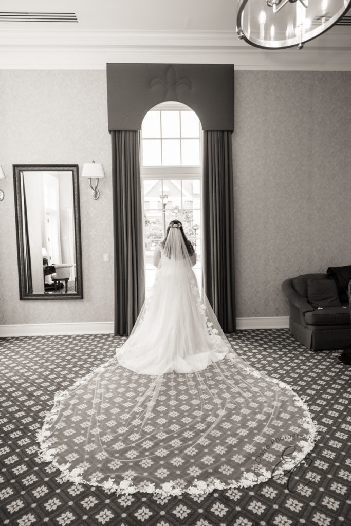 A black and white photo of the back of the bride's dress and veil as she stands in front of a floor to ceiling window.
