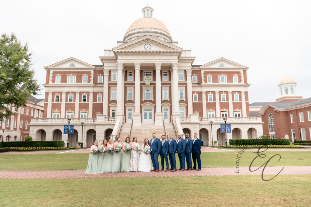 The bride, groom, and wedding party pose for a photo in front of Christopher Newport Hall.