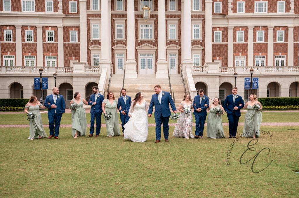 The wedding party walks towards the camera, laughing, in front of Christopher Newport Hall.