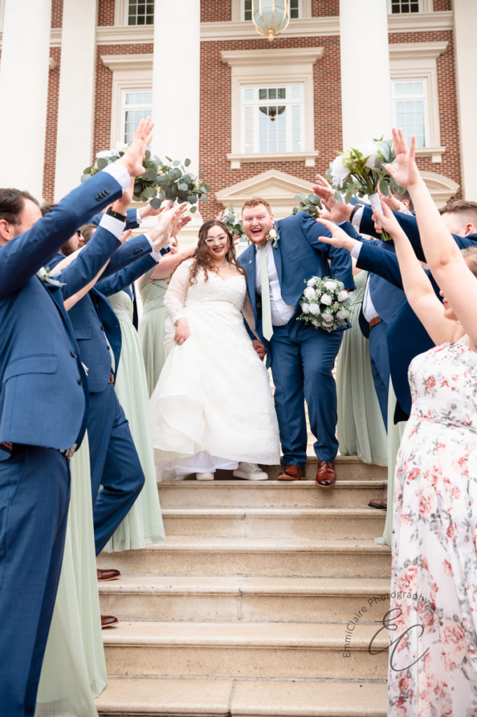 The wedding party stands in front of the church with hands raised while the newlyweds walk through them, smiling.