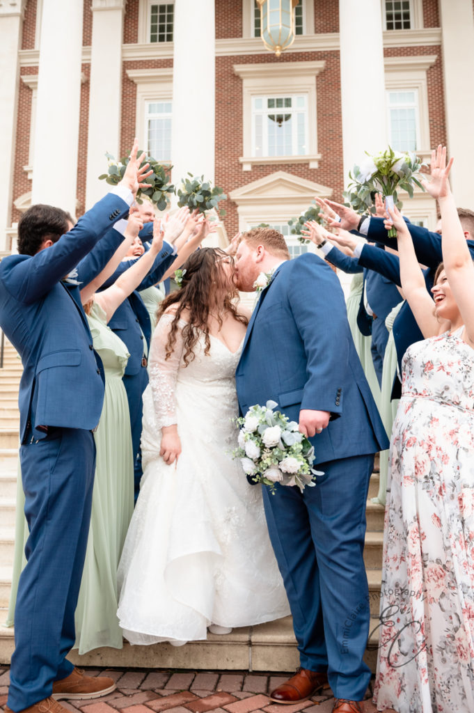 The wedding party stands in front of the church with arms and bouquets raised above their heads in celebration of the newlyweds, who are kissing.