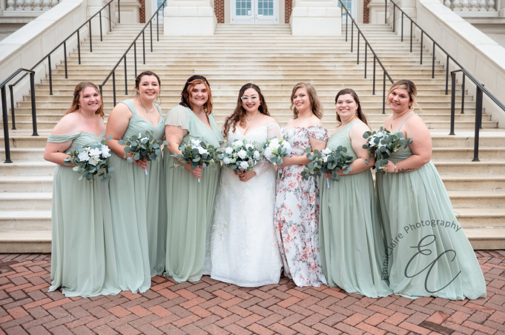 The bride and bridesmaids, clad in shades of soft green, stand in front of Christopher Newport Hall while holding their bouquets.