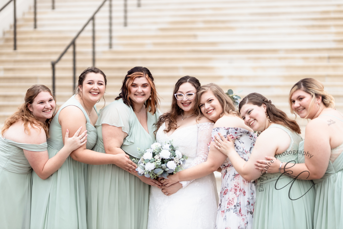 The bridesmaids lean in to snuggle each other and the bride in front of a white stone staircase at Christopher Newport Hall.
