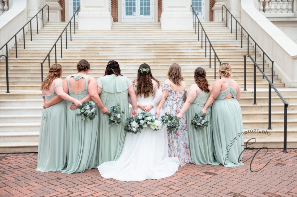 The bride and bridal party stand in front a grand stone staircase at Christopher Newport Hall. Their backs turned to the camera with bouquets held behind them.