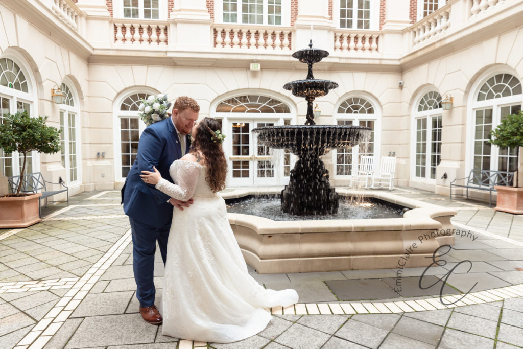 The bride and groom share a kiss in front of a fountain in a heavily windowed/columned courtyard at CNU.