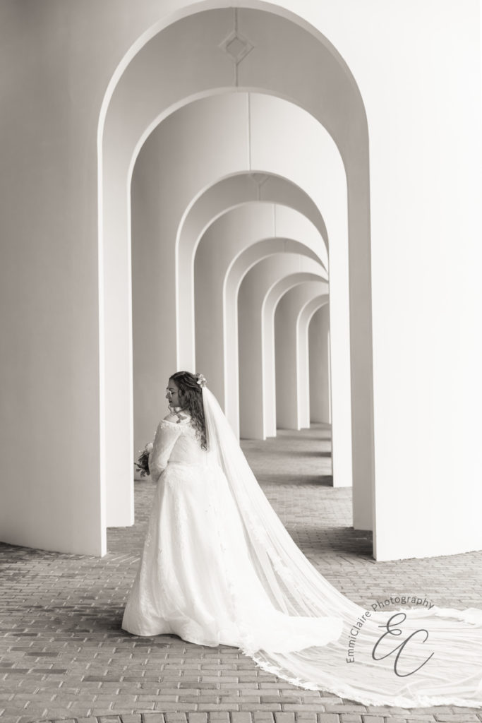 Black and white bridal portrait; the bride has her back turned to the camera, veil flowing behind her beneath arched portico on CNU campus.