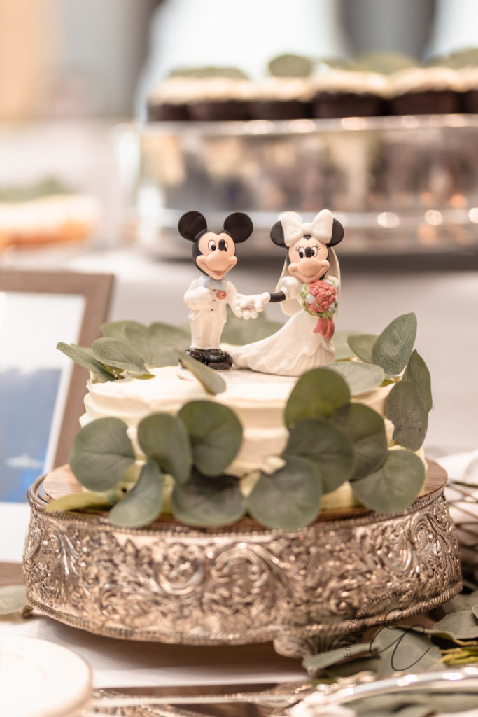 Simple one tiered ruffled wedding cake on silver cake stand, adorned with eucalyptus greenery and Mickey and Minnie Mouse cake topper.