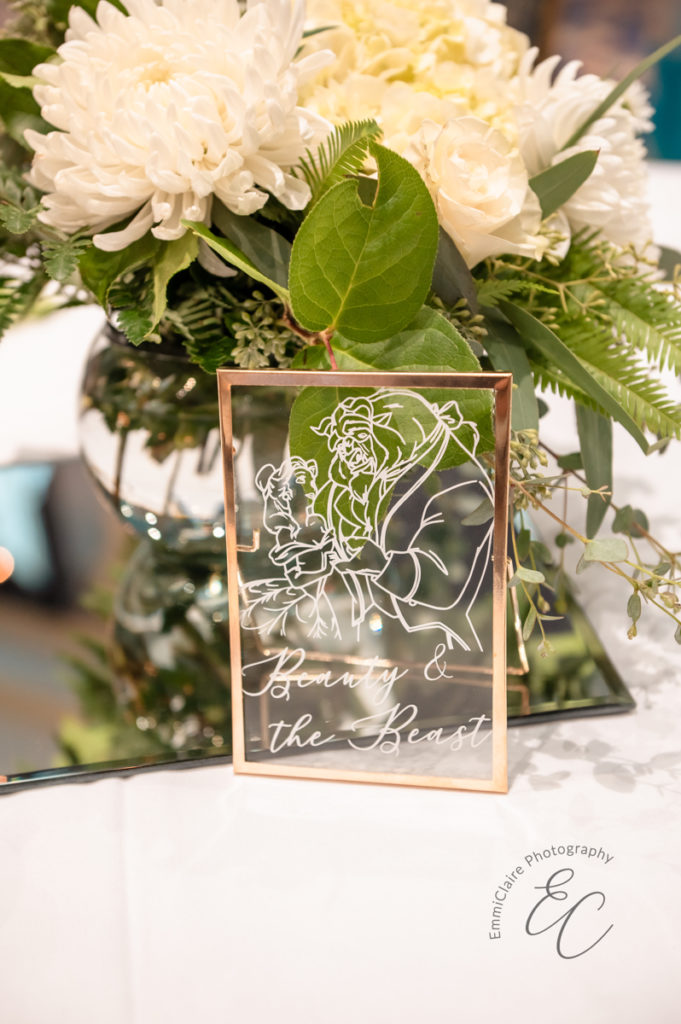 A gold framed glass table name with a DIY drawing of Beauty & the Beast sit in front of a reception centerpiece.