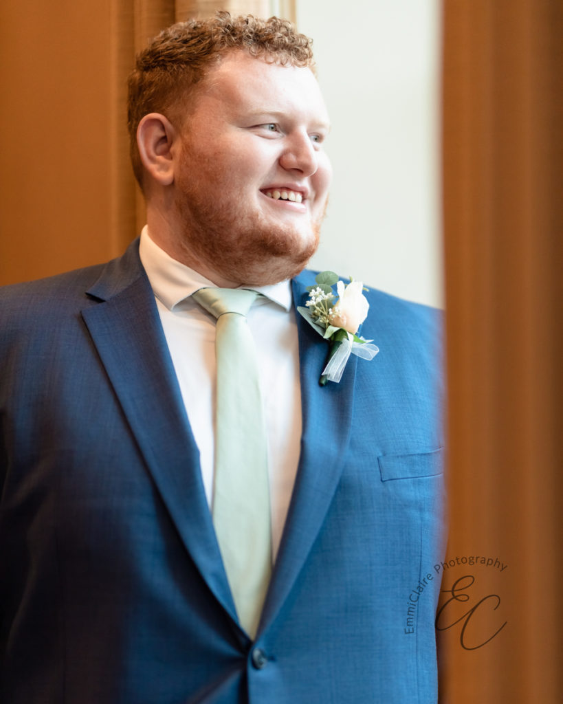 The groom, in a blue suit, smiles as he stares out of a window.