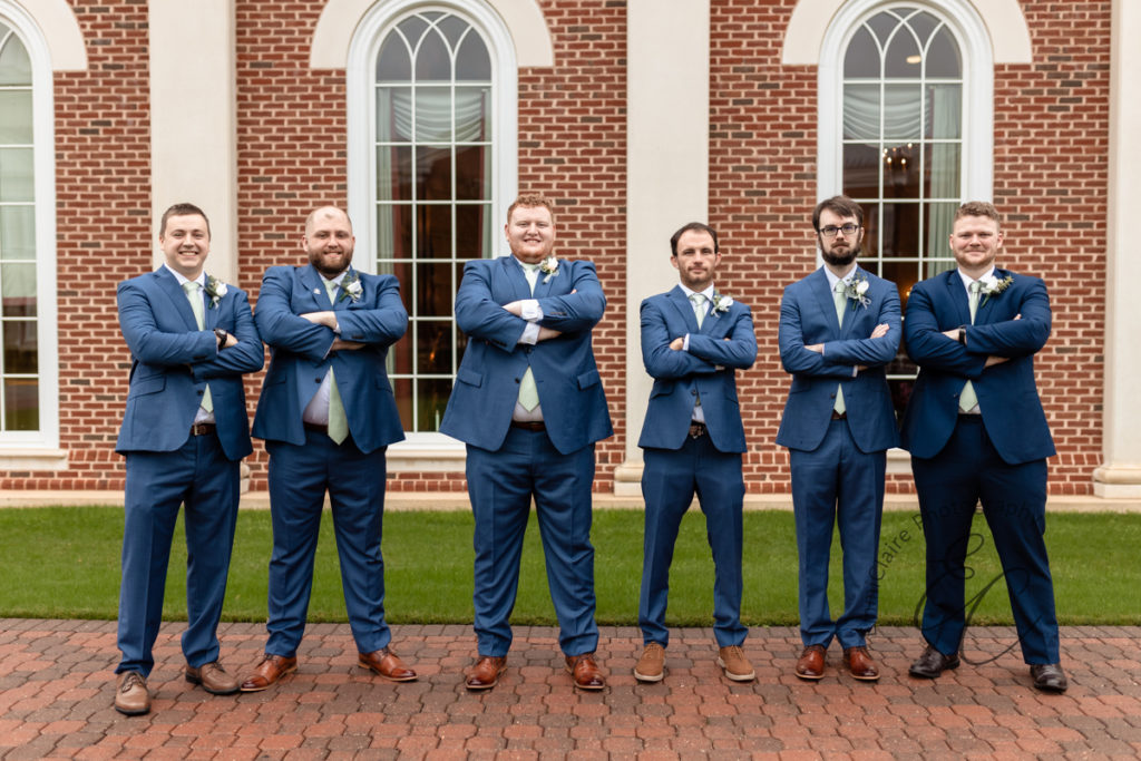 The groom and his groomsmen stand together arms crossed in front of a brick building