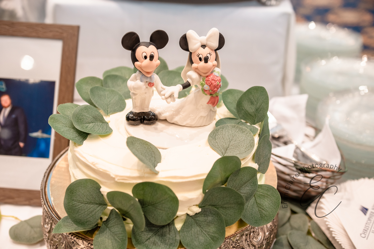 A close up shot of a simple, white ruffled-wedding cake adorned with eucalyptus greenery and topped with a Mickey and Minnie Mouse cake topper.