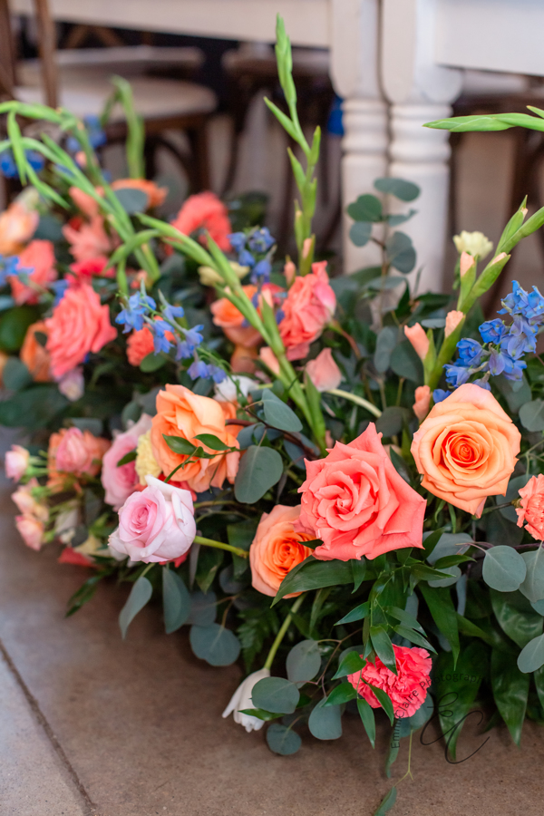 Coral, peach, pink, and blue blooms cascade over lush sage eucalyptus greenery.