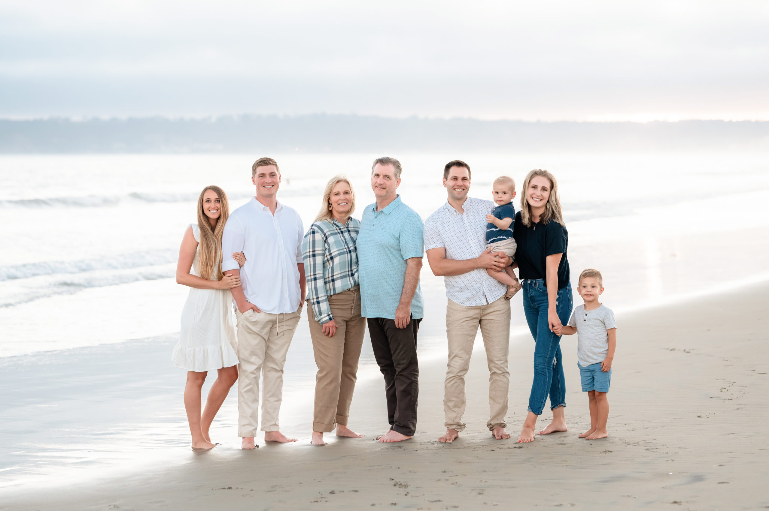 a large family stands together in front of the ocean during their surprise family portrait session at Coronado beach in San Diego, CA.