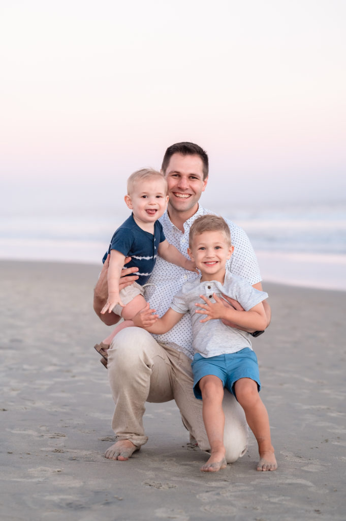 A dad holds his two smiling toddler sons, all smiling during a surprise family portrait session at Coronado Beach in San Diego, CA.